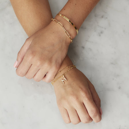 model hands overlapping. on one wrist model wears a gold paperclip bracelet and a gold snake chain bracelet. on other hand model wears a plain gold bead bracelet and a gold letter initial charm on a gold figaro bracelet